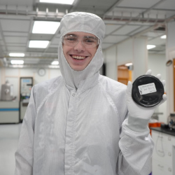 A TST-BOCES high school senior is in the CNF cleanroom, wearing a cleanroom suit, smiling, and holding up a wafer